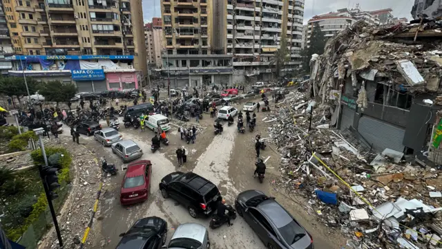 A wide landscape shot of a junction in Beirut, with cars and people gathered in the middle, and damaged buildings in piles of rubble on the right