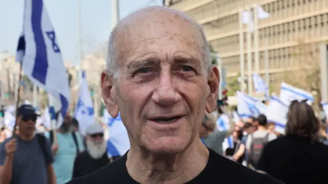 A file photo of Ehud Olmert, standing in a street during a demonstration, with Israeli flags being held up behind him