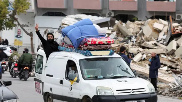 A man holds two fingers up in a victory sign as he hangs from the back of a small van which is driving with luggage strapped to the top through streets lined with debris