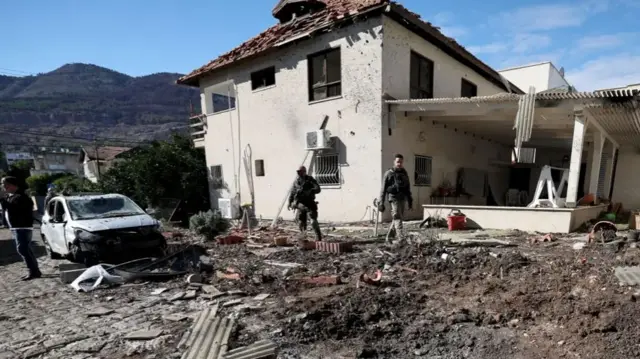 Soldiers stand outside of a damaged home. A car outside is also badly damaged, with the windows smashed