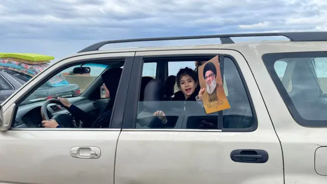 A little girl in the rear seat of a car holds a photograph of a Hezbollah senior leader out the window.