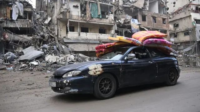 A car sits in the middle of the road in Dahieh. It has mattresses on top, and is in front of a residential street with crumbling buildings