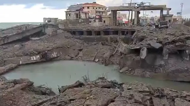 A crater filled with water near partially destroyed buildings.