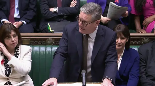 Keir Starmer at the dispatch box speaking during PMQs. He's standing in dark blue suit, white shirt and brown tie, Chancellor Rachel Reeves in blue suit sitting on benches behind him