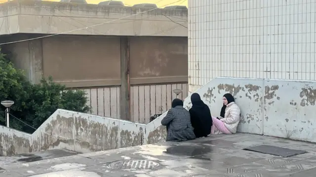 Three women sitting alongside each other at the top of a set of stairs, the picture is taken from a side and behind angle, it looks like it has rained recently as the floor is wet