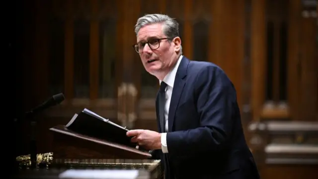 Keir Starmer stands at lectern in House of Commons wearing dark suit, white shirt and black tie. He's holding a folder of documents, the background is a blurred view of the Commons' wooden interior