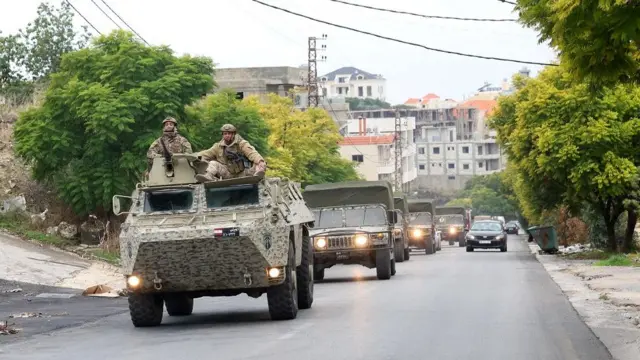 A convoy of Lebanese army vehicles drive down a street. Two soldiers sit out the open top of the one at the front.