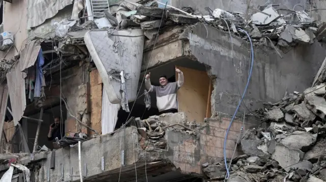 A man stands amidst the ruins of damaged building in southern Lebanon. The floor is above ground, and the man is visible through a hole in the building - he gives two thumbs-up to camera