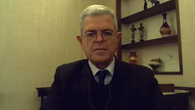 Khalil Helou wearing a suit jacket and tie sitting in a room with beige walls and shelves behind him.