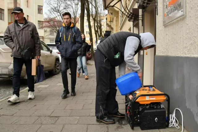 A man wearing a hoodie pours a blue container with petrol into a generator placed beside the wall of a building on a street in Lviv. Passersby walk along the street behind him.