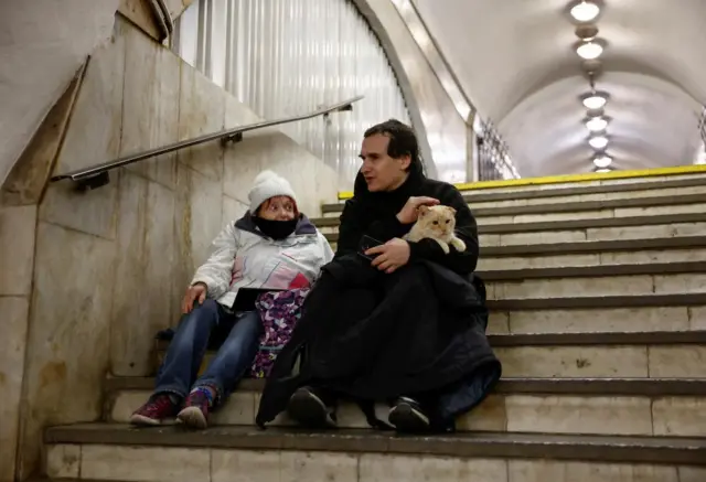 Two people sitting on steps of underground train station in Kyiv, one holding a cat, both wearing winter gear