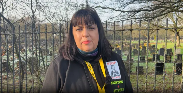 Lee-Bernadette Walford standing in front of a fence next to a cemetery. She has long dark hair and is wearing a black hooded top that says "stop the stink".