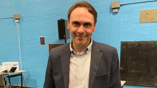 Simon Tagg standing in a hall with a blue wall behind him. He has short brown hair and is wearing a striped shirt and a suit jacket.