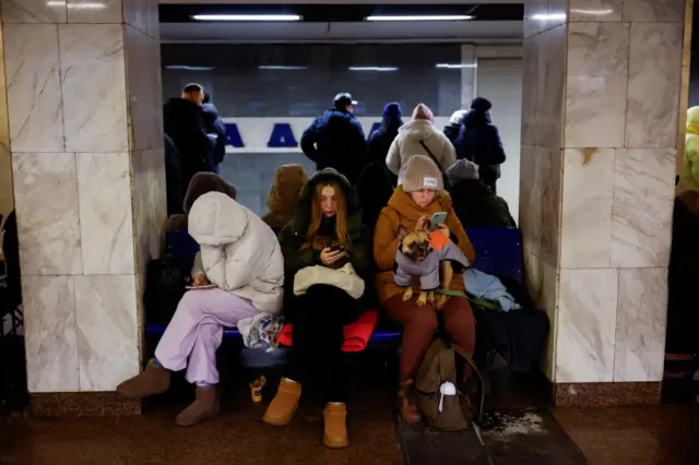 Group of people wearing winter jackets and beanies, sitting in an underground station in Kyiv, looking at their phones. One has a dog on her lap.