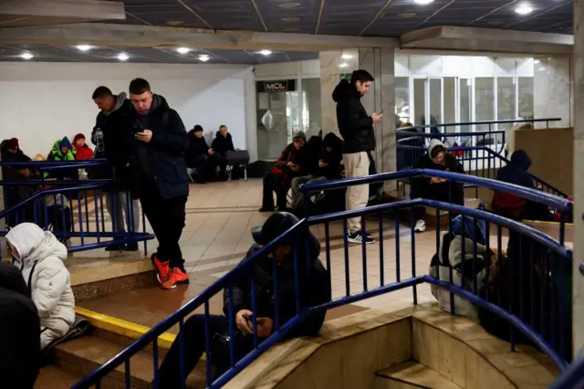 Group of people, many wearing winter gear and looking at their phones, sitting in an underground station in Kyiv