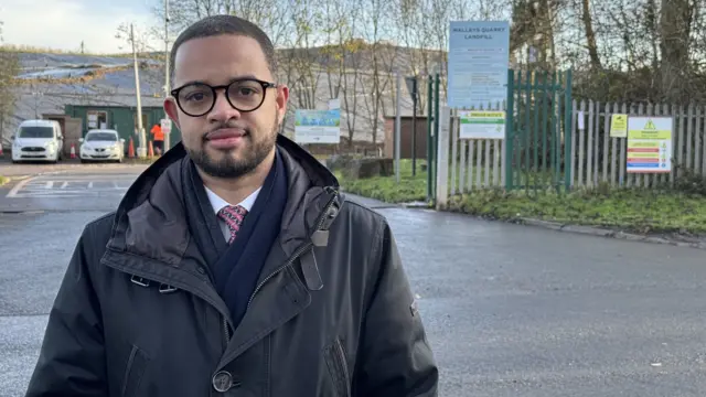 Adam Jogee standing outside Walleys Quarry. He is wearing glasses, a black coat, a black scarf, a white shirt and a tie.