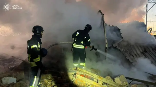 A firefighter in Lutsk, Volyn in northwest Ukraine, holds a hose which is spraying water on a fire underneath a collapsed building. Another firefighter holds up the hose behind him. Both standing on rubble with lots of smoke rising from the fire.