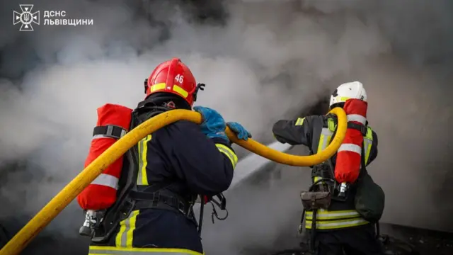 Two firefighters in full gear hold a yellow hose, both wearing helmets and red extinguishers on their backs, with water coming out of the hose and smoke covering the entire background of the image