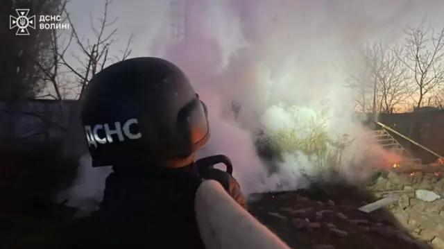 Early morning scene showing the back of a person in a black helmet with Ukrainian writing and lots of smoke rising from a fire with bare trees in the background