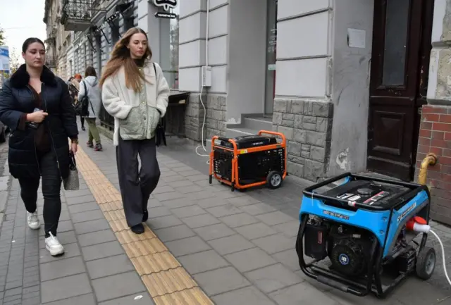 Two people walk past two portable generators on a street, plugged into power points in the external walls of buildings