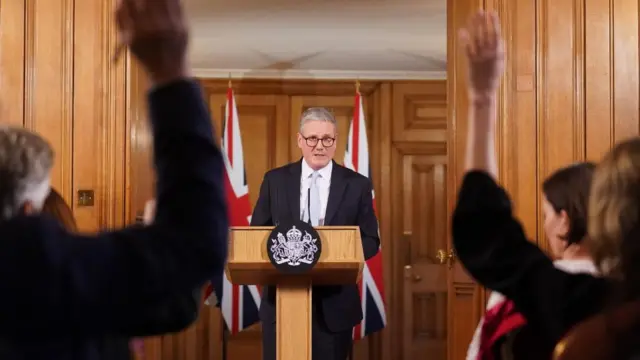 Sir Keir Starmer stands at a podium in front of two Union flags, as people seated in front of him raise their hands