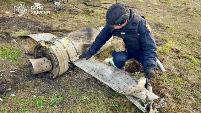 A uniformed worker with a helmet and padded vest inspects a piece of a missile on a grassy area in Ukraine