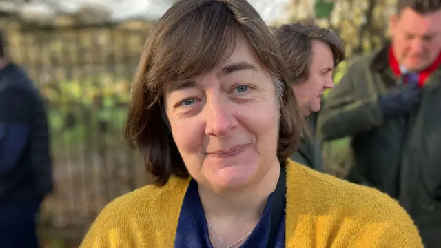 A woman with mid-length brown hair wearing a yellow cardigan, blue top.