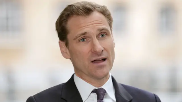 Chrs Philp talking to someone who is just beyond the camera. He has blue eyes and light brown hair. He is wearing a black suit and tie.