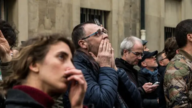 A crowd of onlookers hold their hands to their faces with devastated expressions