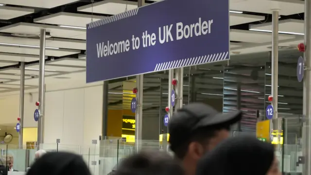 A sign saying Welcome to the UK Border. It is hung above a queue of people and has a dark blue background and white lettering.