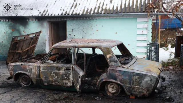 A completely burnt out car with one of the doors open, the front hood up, no windows, no tyres on the wheels. Building behind car shows fire damage