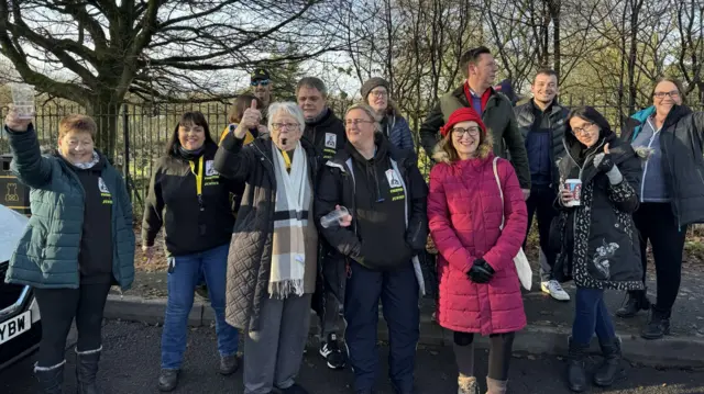 A number of people outside the cemetery next to the Walleys Quarry landfill site. Some are holding their thumbs and hands up, while one is seen blowing a whistle.