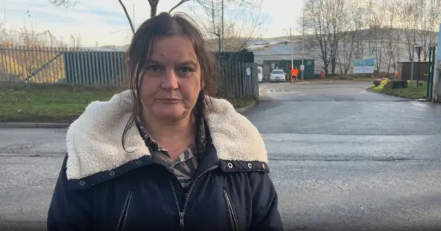 A woman wearing a black coat and a white fluffy hood lining stands outside the entrance to Walleys Quarry landfill site