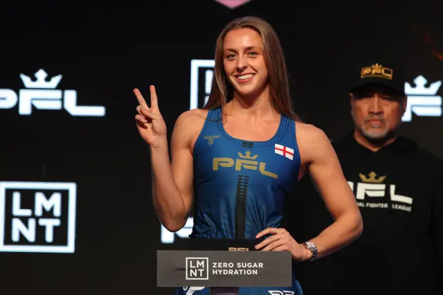 Dakota Ditcheva puts up two fingers in a peace sign as she stands on the scales