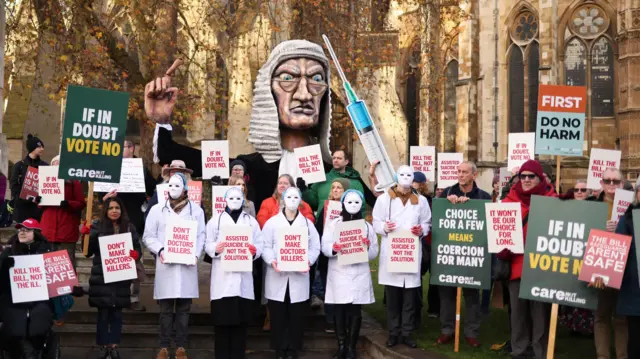 Demonstrators dressed in white medical coats hold posters that read "Don't make doctors killers"; behind them is a large model of a judge in a white wig