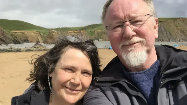 Mary Maycroft and her bearded husband stand smiling on a beach