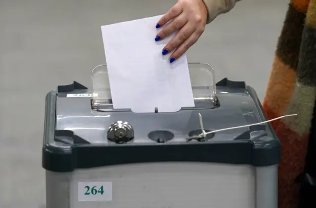 A grey ballot box sits in the middle of the image. A hand with painted blue nails is dropping a white piece of paper into a slot at the top of the box.