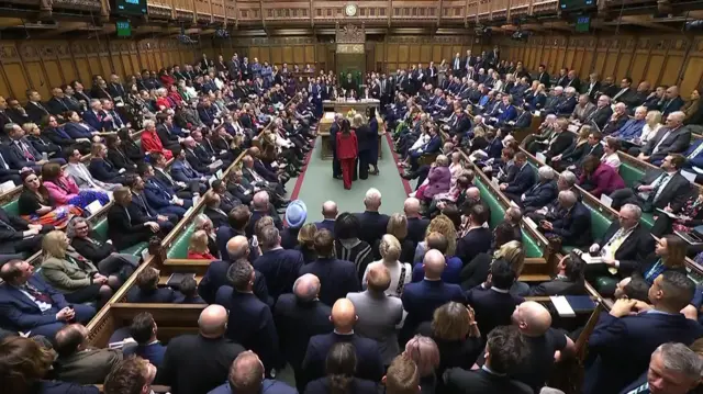 The House of Commons with benches on other side of the middle aisle. Seats are full of MPs with many standing up in the aisle so they can see the results be announced