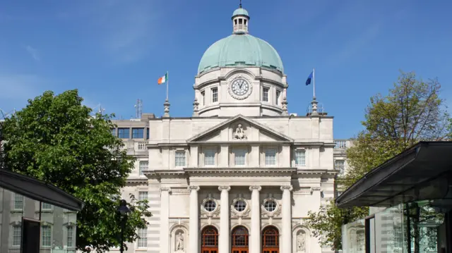 A large white building sits against a bright blue sky. On either side are modern glass fronted buildings.