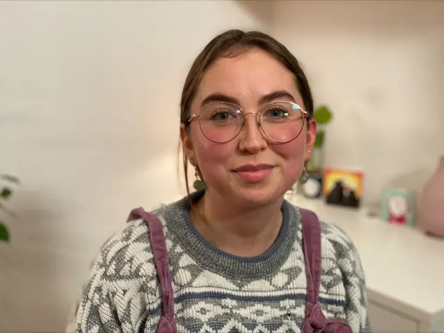 Maddie Cowey is shown in her home. She is wearing a knit jumper and a pair of dungarees, and gold-rimmed round glasses.