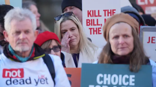 Woman wipes tears from her eyes among a crowd of demonstrators. T-shirts reading 'not dead yet' can be seen, as well as a number of different placards