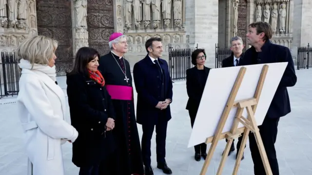 Macron is talking to Anne Hidalgo, the mayor of Paris. Macron is wearing a navy coat and suit and tie. Hidalgo has a black coat on and orange scarf.