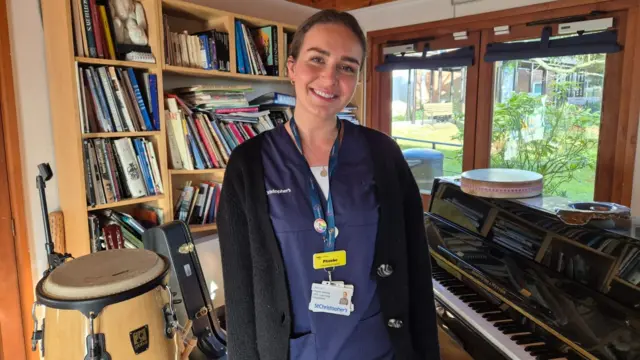 Nurse Phoebe stands in a home in front of a piano and a drum. There is a bookshelf and a window behind her.