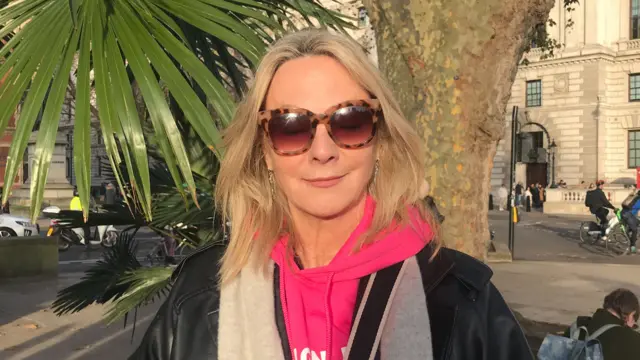 Woman wearing pink hoodie, scarf and leather jacket looks into the camera. She's smiling slightly and wearing sunglasses.