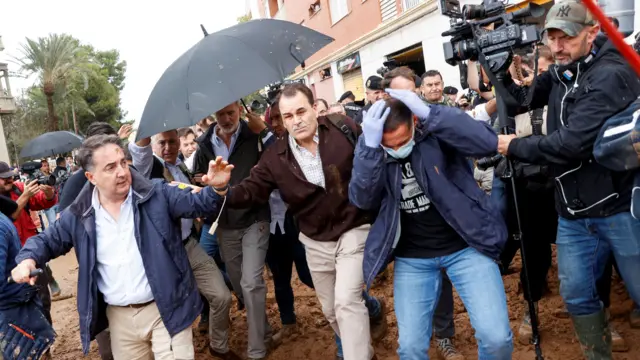 Three men stand in line in front of the king, reacting to mud being thrown. The king is behind them, partially protected by an open umbrella