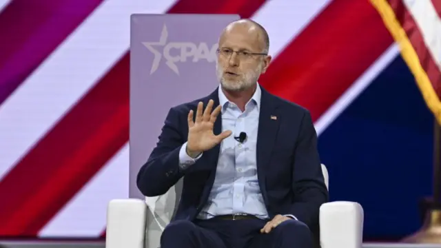 Brendan Carr, in an unbuttoned blue suit and white shirt, gestures with his right hand while seated at a Conservative Political Action Conference panel