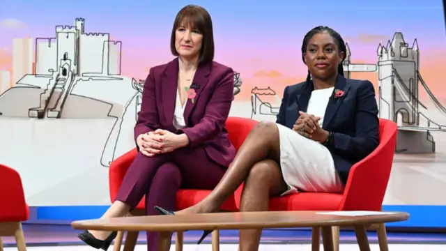 Rachel Reeves in plum suit and white blouse sitting on red chair. To her left is Kemi Badenoch in black jacket and white dress also sitting. Both are wearing a poppy pin