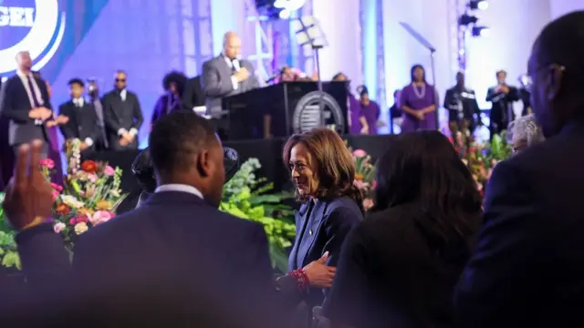 Harris smiles as she greets a churchgoer in the foreground - in the background a minister gives a speech with a choir behind him