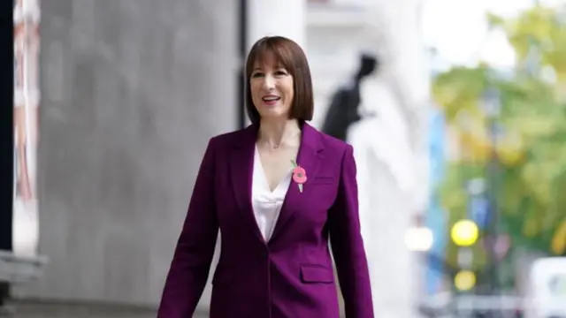 Rachel Reeves entering New Broadcasting House on Sunday morning. Shot is cut just below the waist, Reeves is wearing a plum suit jacket with a white blouse. Pinned to the left lapel is a poppy pin