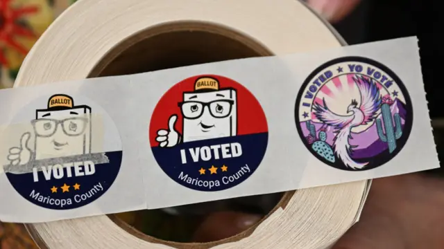 Three round stickers are shown on a strip of paper. Two feature a cartoon ballot with a baseball hat that is giving a thumbs up. The third features catci, desert plants and a phoenix. All three have text that says "I voted, Maricopa County"